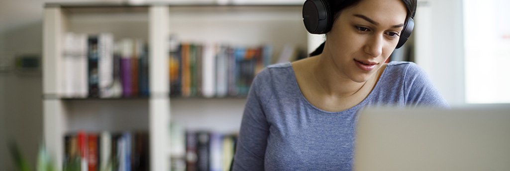 Une femme assise, un casque audio sur les oreilles, regarde son écran d'ordinateur portable et prend des notes en même temps.