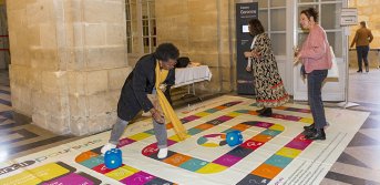 Des personnes en train de jouer au jeu de société de l'Agefiph Handipoursuite, version géante avec le plateau de jeu à même le sol, sur lequel les personnes marchent directement.