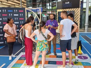 Romain de face avec un polo Agefiph violet anime le jeu handipoursuite pour un groupe de 4 personnes, 2 femmes sur sa droite et 2 hommes sur sa gauche