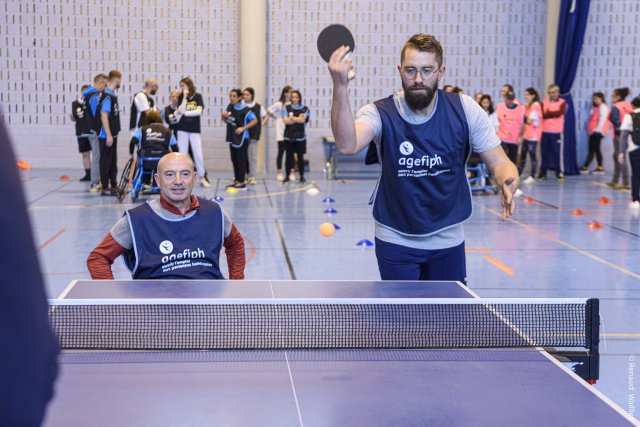 Deux participants s'exercent au tennis de table en double. L'un des deux est assis dans un fauteuil.
