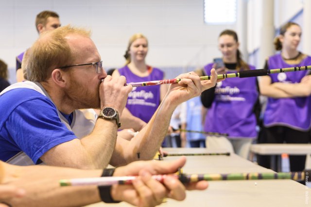 Un joueur de l'équipe violette se prête au défi de la sarbacane qu'il porte à sa bouche pour viser la cible