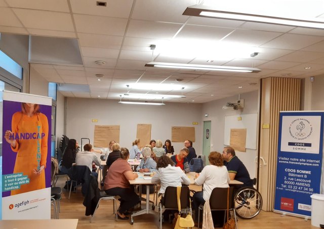 Des référents handicap réunis sur 3 tables espacées travaillent en sous-groupe sur un atelier. A gauche de la photo, se trouve un roll'up Agefiph représentant une femme tenant l'inscription Talent Handicap. A droite de l'image un roll up du Comité Départemental Olympique et Sportif est positionné. Il est de couleur bleu et blanche avec le logo représentant les anneaux et un coq et l'adresse.