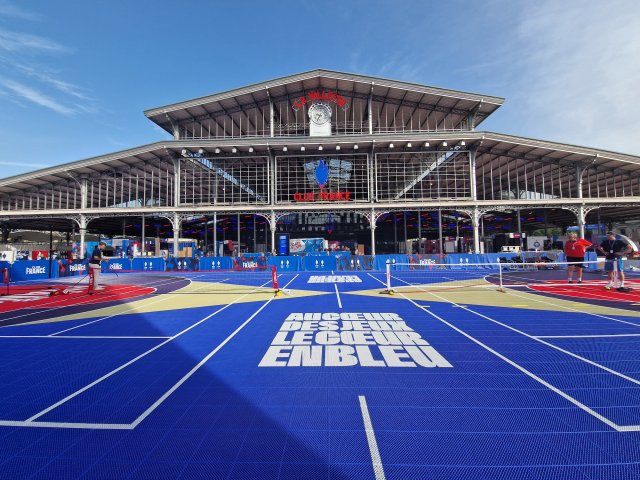 Plan large de la hallle de la villette au couleur du Club France. Au premier plan se trouve un terrain multisport bleu avec une inscirption au sol en majuscules et en blanc : "Au cœur des Jeux, le cœur en bleu"