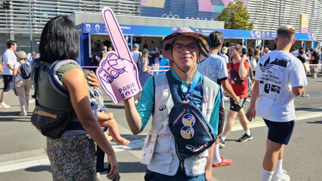 Michel au centre de l'image porte une main géante pour indiquer le chemin à suivre aux spectateurs. Il a derrière lui le Stade Pierre Mauroy
