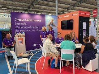 Des personnes sont présentes sur le stand de l'Agefiph violet et orange. 5 personnes sont assises en premier plan en train de jouer à un quizz. Stéphanie et une collègue au second plan sourient pour la photo. Une autre personne consulte un ordinateur sur la gauche de l'image.