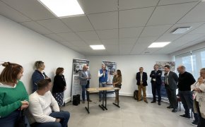 Au centre de l'image, une personnalité vêtue d'un doudoune sans manches bleue prend la parole au micro devant deux kakémonos. Il est accompagné d'une femme à sa gauche et d'un homme à sa droite. De chaque côté 9 personnes, 5 à droite et 4 gauche de l'image écoutent son discours.