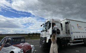 Jérémy Soots fait une démonstration de la plateforme élévatrice lui permettant d'accéder à la cabine de son camion. Une photographe se tient devant pour immortaliser la scène. Sur la gauche de l'image se trouve une 2 chevaux violette, blanche et bleu portant les logos de l'Agefiph et l'Aftral sur le capot