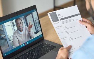 Un homme est assis devant une table sur laquelle est posé son ordinateur portable. Il tient un CV dans ses mains. Il est en train de faire passer un entretien en visio a un autre homme dont on voit le visage sur l'écran d'ordinateur.