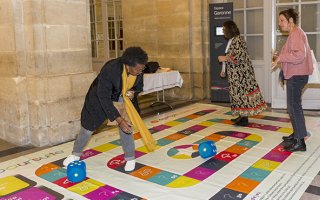 Des personnes en train de jouer au jeu de société de l'Agefiph Handipoursuite, version géante avec le plateau de jeu à même le sol, sur lequel les personnes marchent directement.
