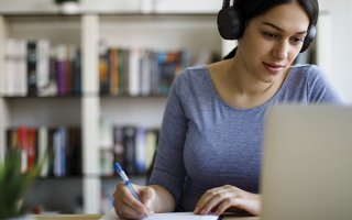 Une femme assise, un casque audio sur les oreilles, regarde son écran d'ordinateur portable et prend des notes en même temps.