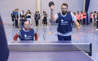 Deux participants s'exercent au tennis de table en double. L'un des deux est assis dans un fauteuil.