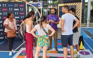 Romain de face avec un polo Agefiph violet anime le jeu handipoursuite pour un groupe de 4 personnes, 2 femmes sur sa droite et 2 hommes sur sa gauche