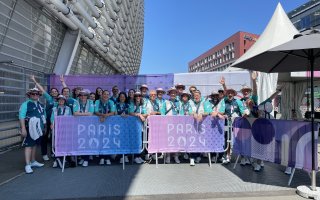 Un groupe de volontaires posent derrière une grille sur laquelle se trouve une bâhce PAris 2024 rose et violette