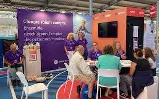 Des personnes sont présentes sur le stand de l'Agefiph violet et orange. 5 personnes sont assises en premier plan en train de jouer à un quizz. Stéphanie et une collègue au second plan sourient pour la photo. Une autre personne consulte un ordinateur sur la gauche de l'image.